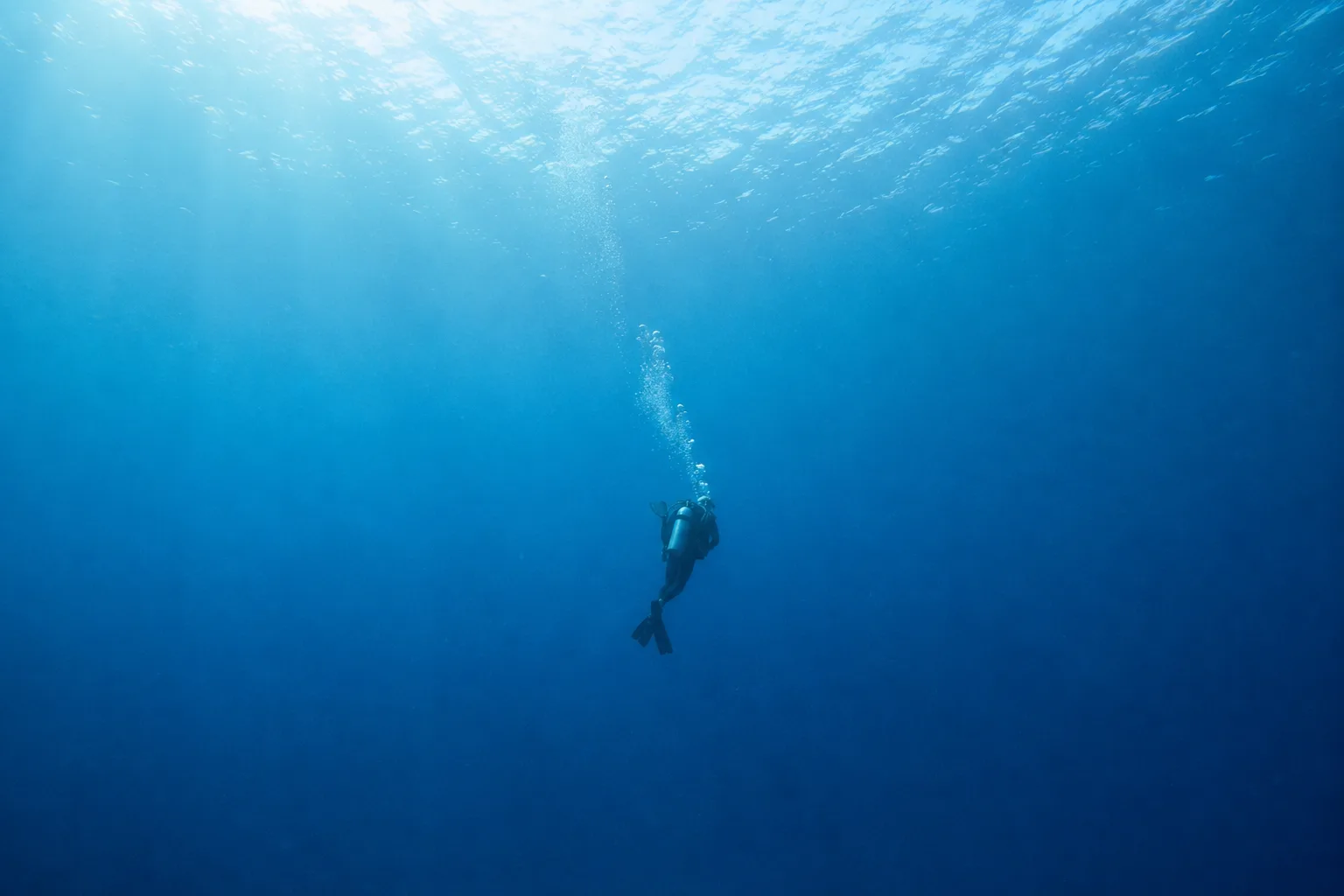 A lone scuba diver descending through clear blue water, sunlight filtering from the surface above